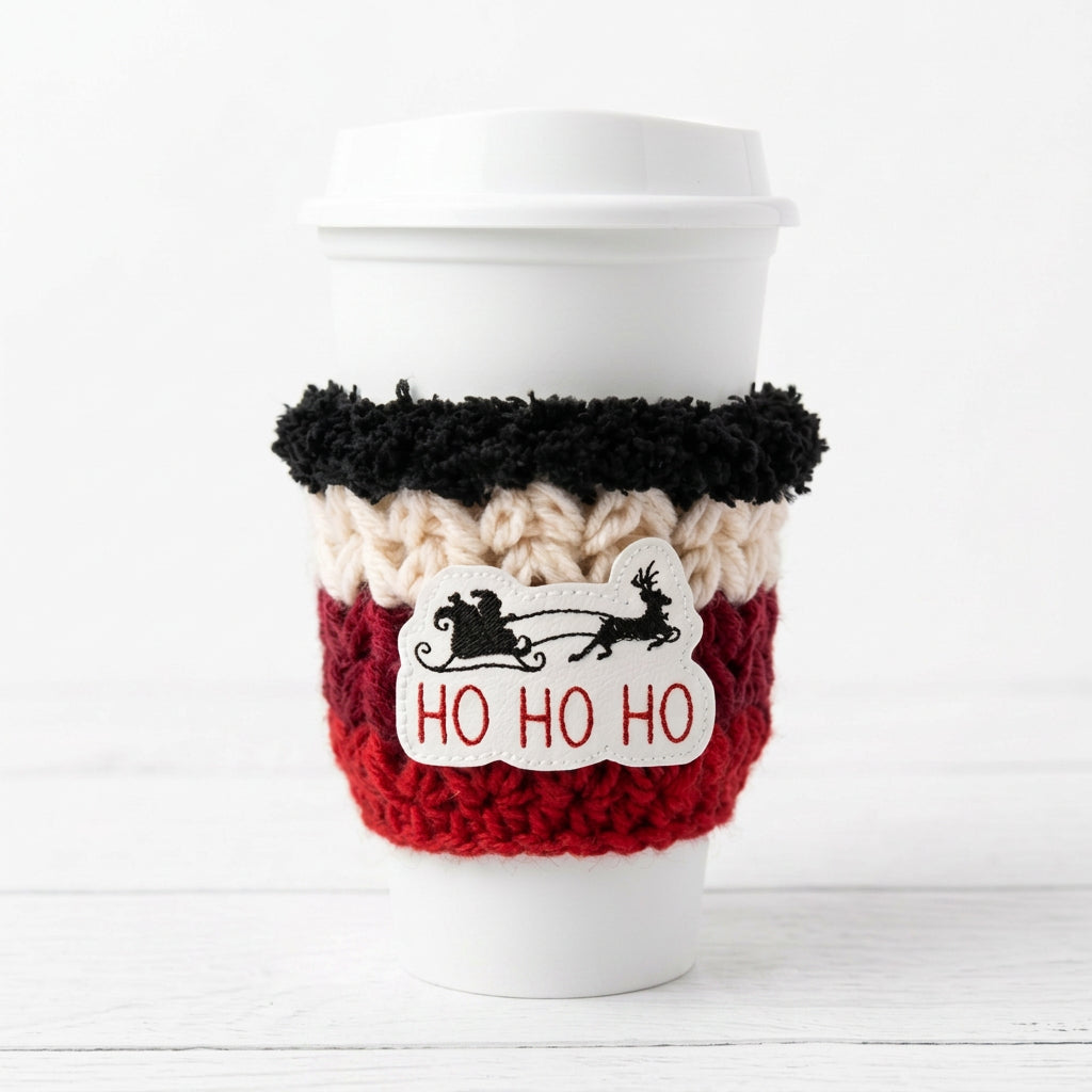 A white paper coffee cup with a white lid, wrapped in a knitted holiday cozie featuring black, cream, and red stripes, with a white patch that reads 'HO HO HO' and shows Santa in a sleigh.