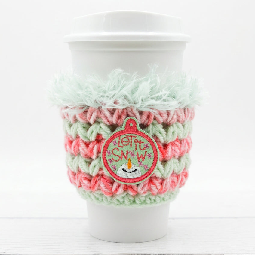 A white paper coffee cup with a white lid, wrapped in a handmade crocheted holiday cozy sleeve featuring pink and mint green stripes, a fuzzy mint green top trim, and a red 'Let It Snow' ornament patch.