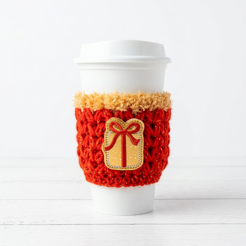 A white disposable coffee cup with a white lid, wrapped in a red and gold knit sleeve featuring a gold gift box patch with a red bow.