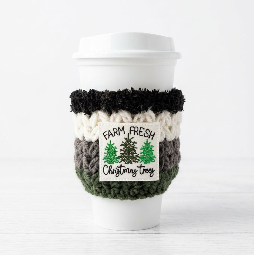 A white reusable coffee cup with a white lid, wrapped in a handmade knit holiday cozy sleeve. The cozy features a black top band, a cream textured middle band, a dark gray lower band, and a white rectangular patch with green Christmas trees and the text 'FARM FRESH Christmas trees'.