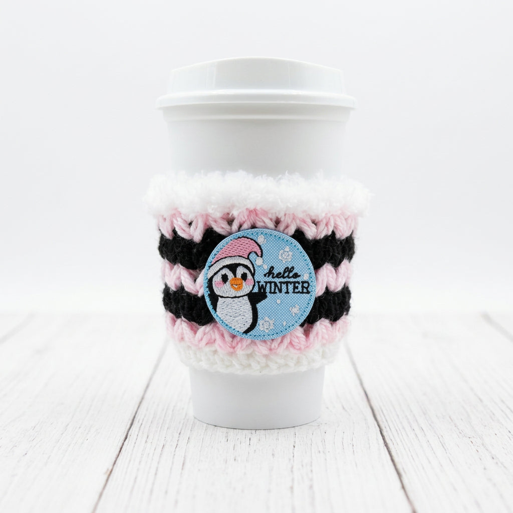 A white disposable coffee cup with a white lid, wrapped in a black and pink striped knit sleeve with fluffy white trim. A round blue patch on the sleeve features a penguin wearing a pink hat and the words 'hello WINTER'.