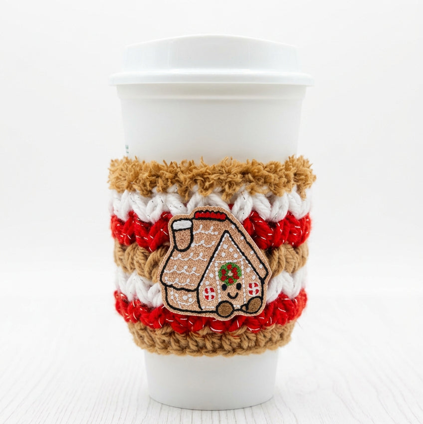 A white paper coffee cup with a white lid, wrapped in a handmade knit holiday cozy sleeve featuring red stripes, a brown jute trim, and a gingerbread house design on the front.